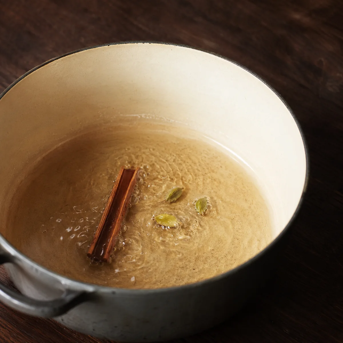 A cinnamon quill and two green cardamom pods sitting in shimmering oil in a heavy cream-enamel pot, about to bloom
