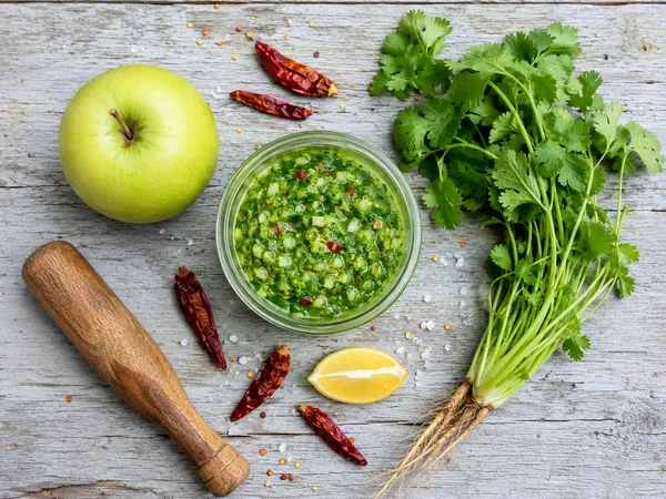 A glass jar of bright green apple and coriander chutney, surrounded by a whole green apple, dried red chillies, fresh coriander with roots, a lemon wedge and a wooden pestle.